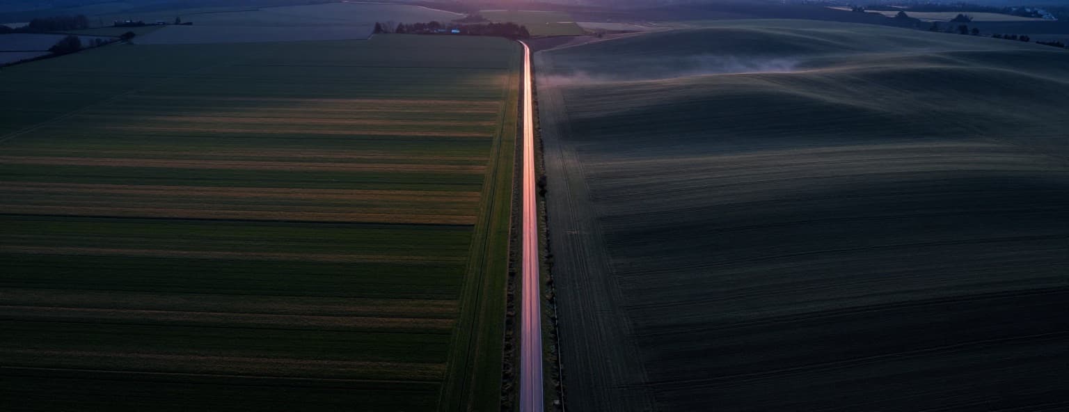 Aerial view at dusk of two differently patterned farmlands meeting along a straight road lit in copper light